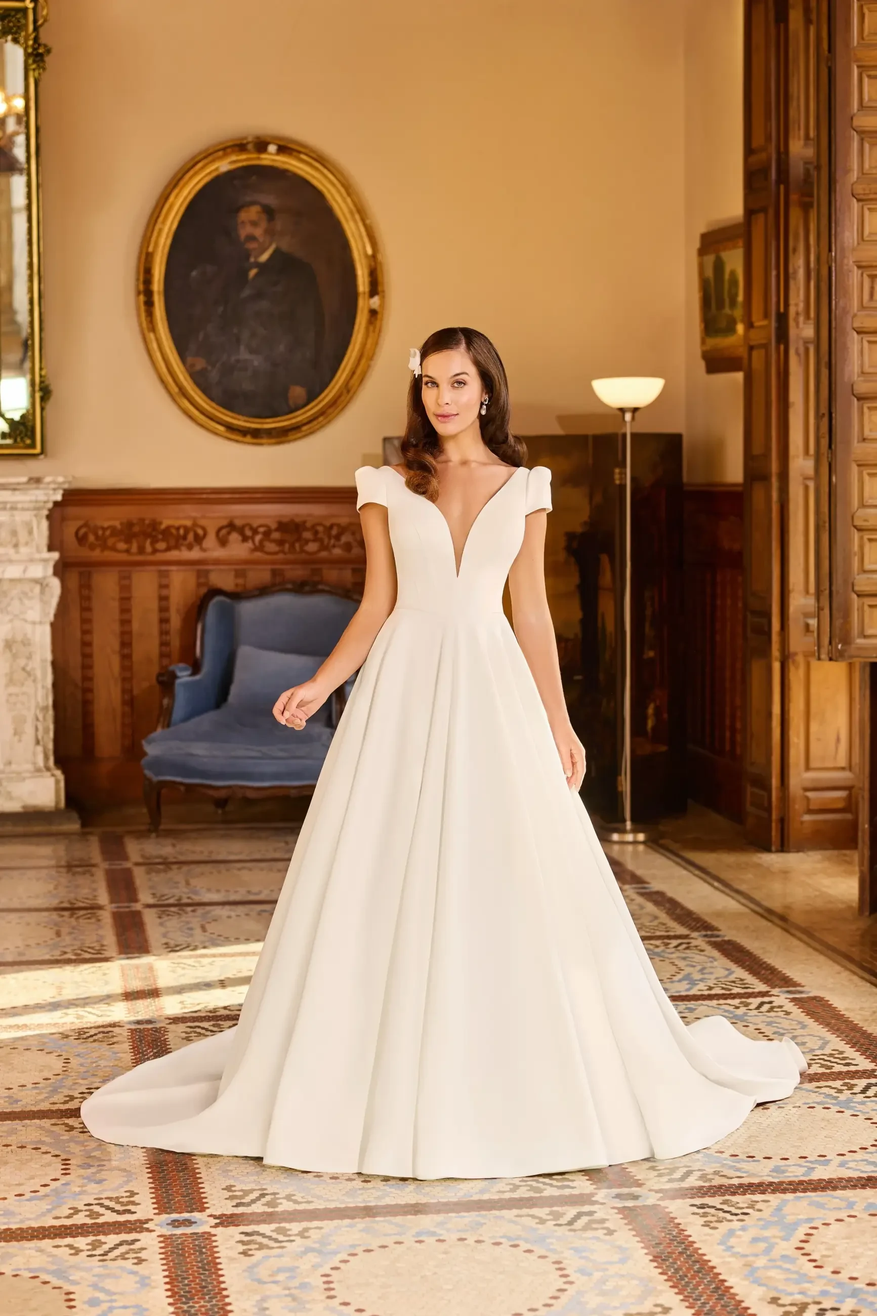 A bride stands in a grand room wearing a white ball gown with a deep V-neck and cap sleeves. The room features wooden details and a portrait on the wall.