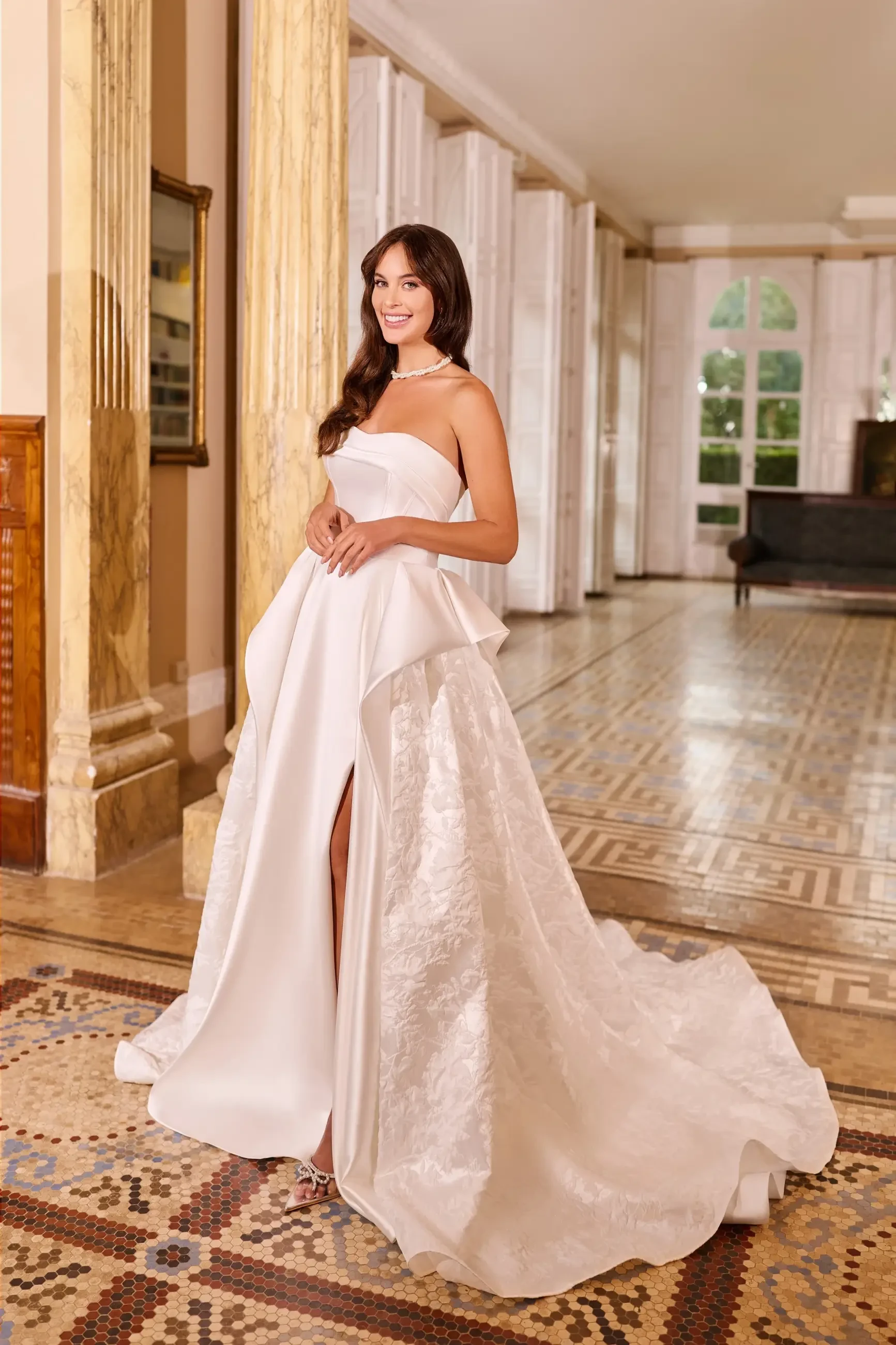 A woman in a strapless white gown with a flowing skirt stands in a grand interior space with large windows and ornate flooring.