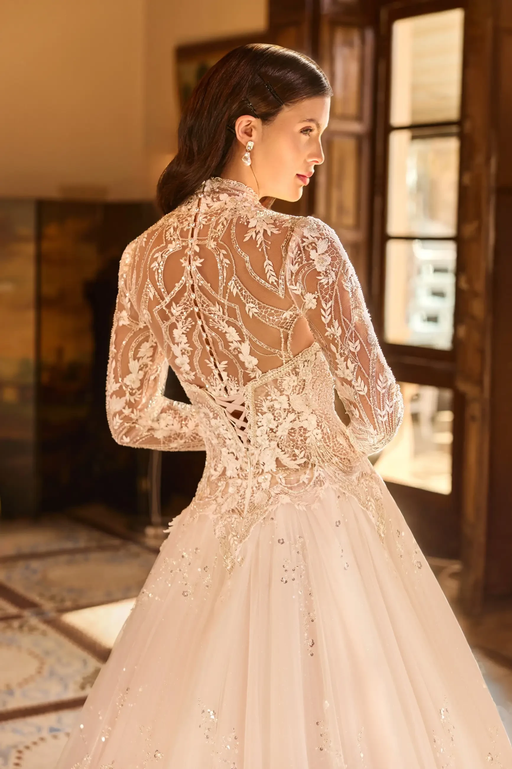A woman wearing a white wedding dress with intricate lace detailing on the back, standing in front of a window with wooden frames.