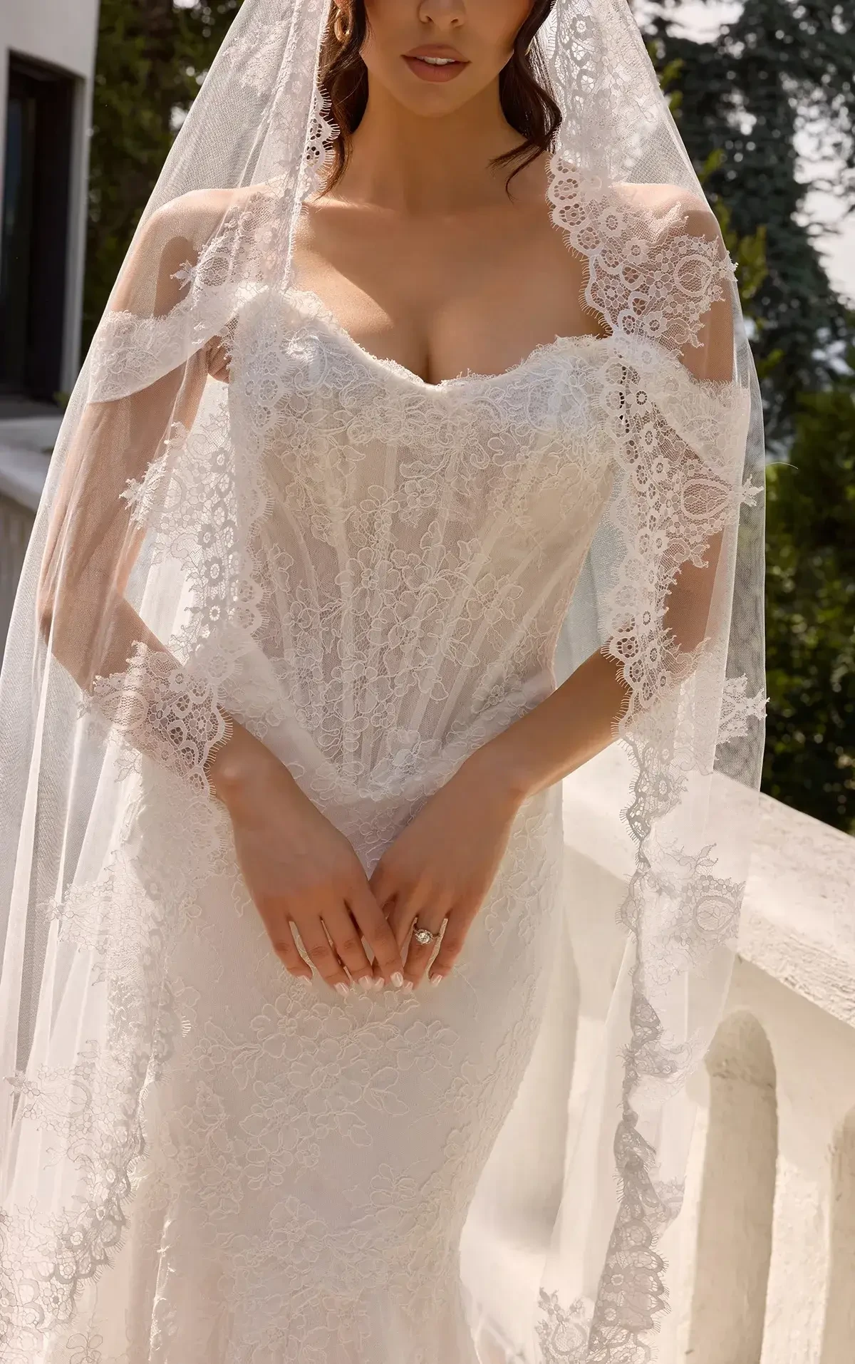 A woman in a white, off-the-shoulder wedding dress with lace details and a long train poses confidently on a wood floor with a neutral background.
