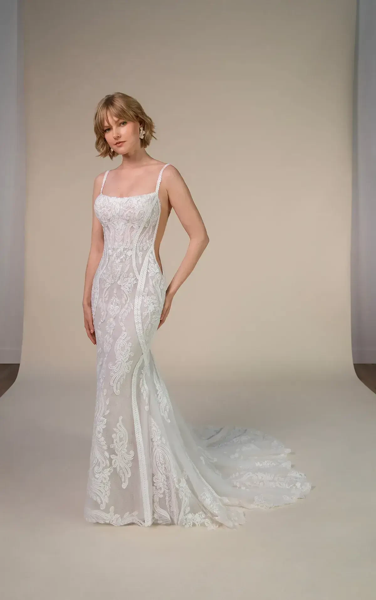 Woman in a form-fitting, lace wedding dress with a train, standing on a neutral backdrop. Her expression is serene, fitting the elegant tone.