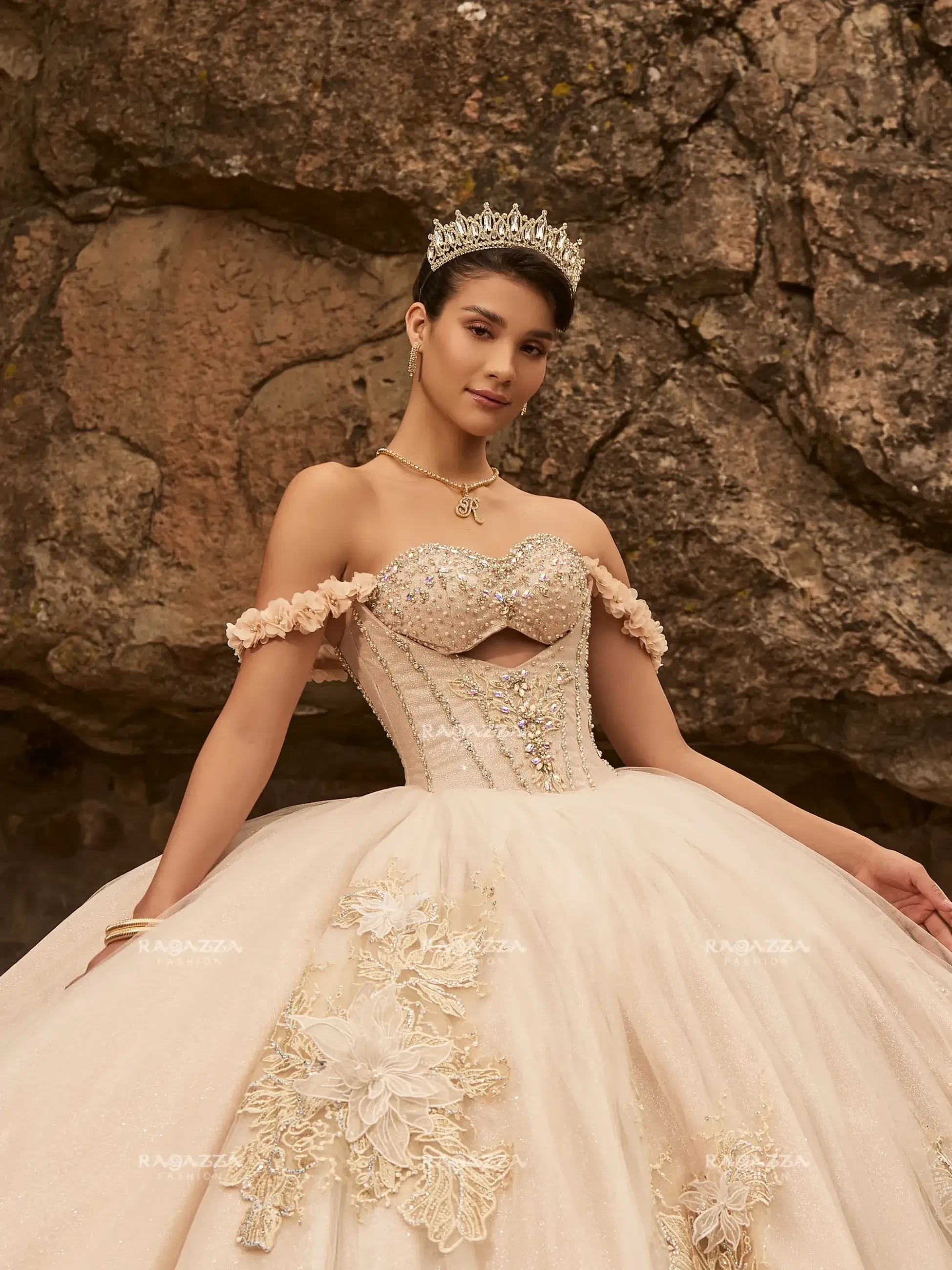 A model wearing an elegant gown with off-the-shoulder sleeves, a fitted bodice adorned with embellishments, and a voluminous skirt, posing against a rocky backdrop. She is also wearing a tiara.