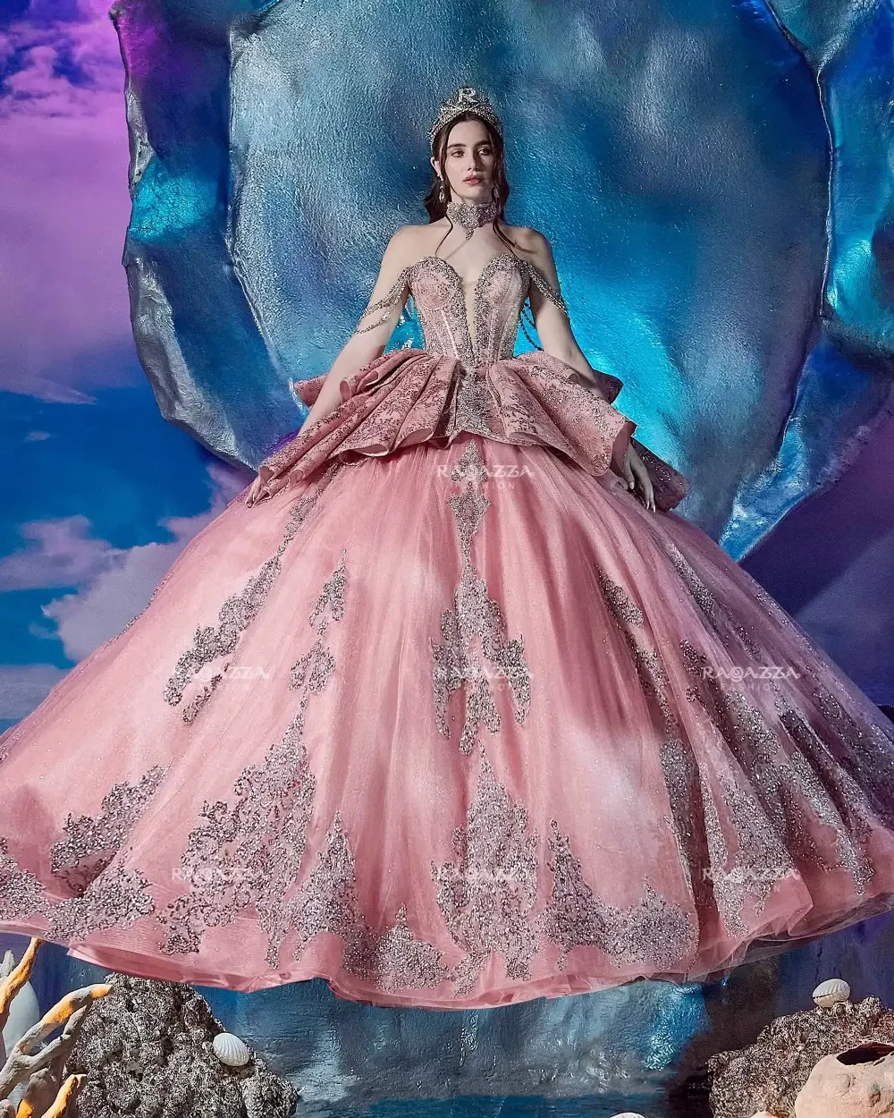 A woman in an elaborate pink gown with silver embellishments, posing in front of a vibrant blue and purple background.