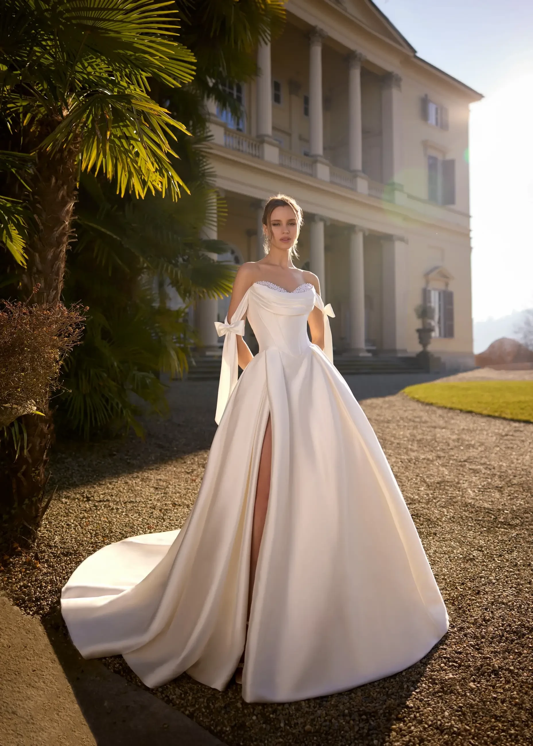 Elegant woman in a flowing white gown stands outdoors, highlighted by sunlight. She poses gracefully in front of a grand mansion, surrounded by lush greenery.