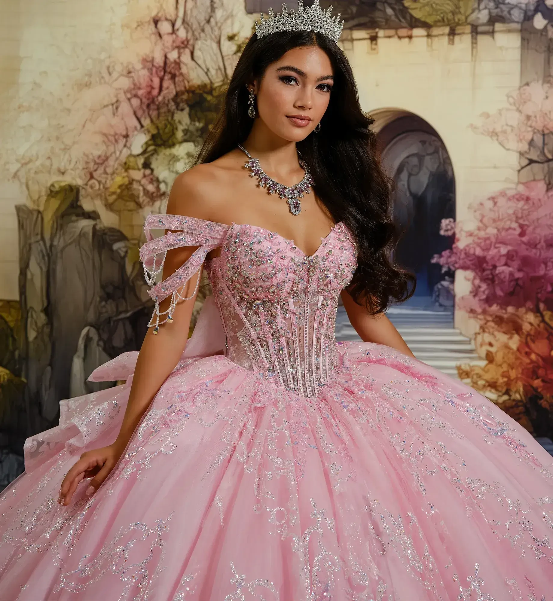 A person wearing a pink ball gown with sparkling details and a tiara, posing in front of a pastel-colored backdrop.