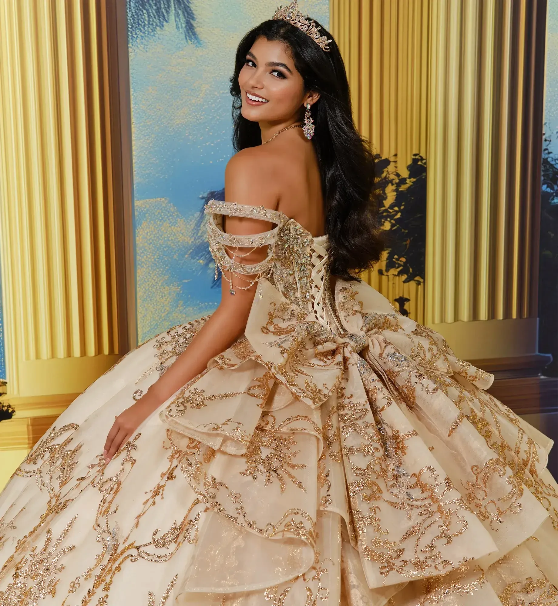 A young woman wearing an elegant, embellished gown with a full skirt, smiling and posing with her back towards the camera. She has long, dark hair and is adorned with jewelry and a tiara, set against a backdrop featuring golden columns.